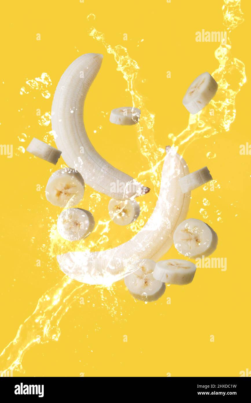 Several bananas and banana slices suspended in the air and surrounded by fresh water on a yellow background Stock Photo