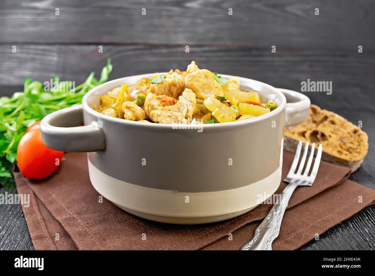 Stew of chicken breast, tomatoes, stalked celery, carrots, green peas and onions in a tureen on a napkin on black wooden board background Stock Photo