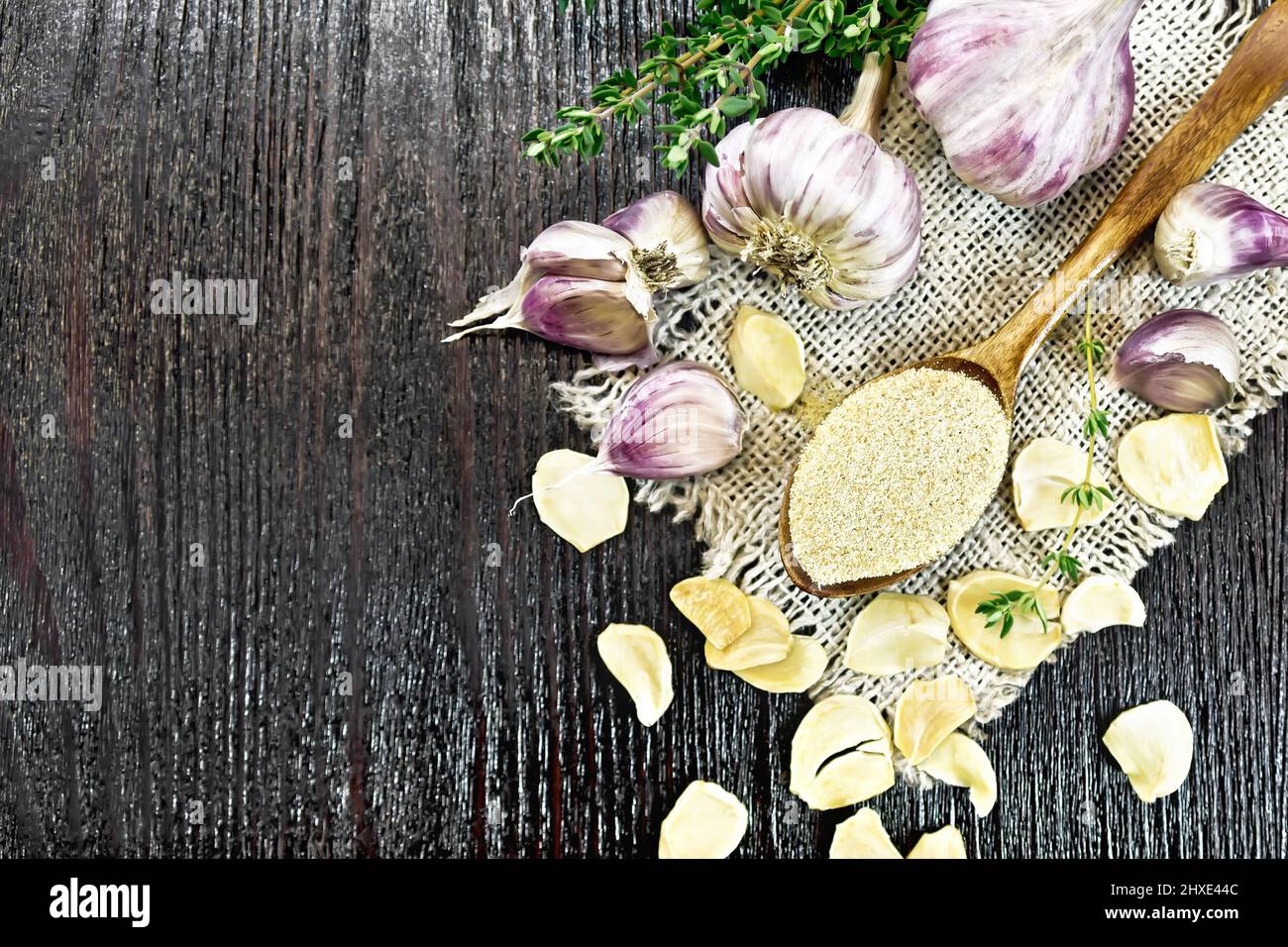 Ground garlic in a spoon on a burlap, dried and fresh garlic, bunch of thyme on background of wooden board from above Stock Photo