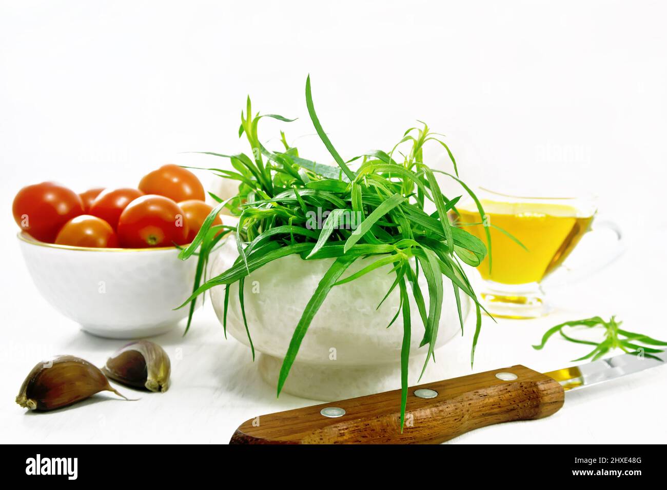 Fresh tarragon in a mortar, tomatoes and champignons in bowls, vegetable oil in gravy boat, garlic cloves and a knife on light wooden board background Stock Photo