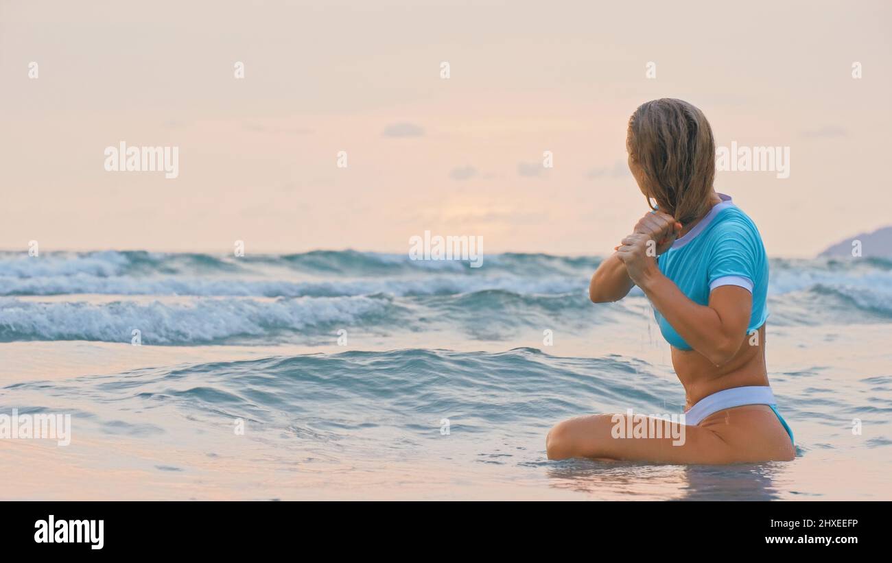 SLOW MOTION GLOW MIST GOLD SUNSET CINEMATIC VIEW: Attractive woman sitting on water sea. Girl in turquoise swimsuit. Freedom paradise holiday vacation Stock Photo