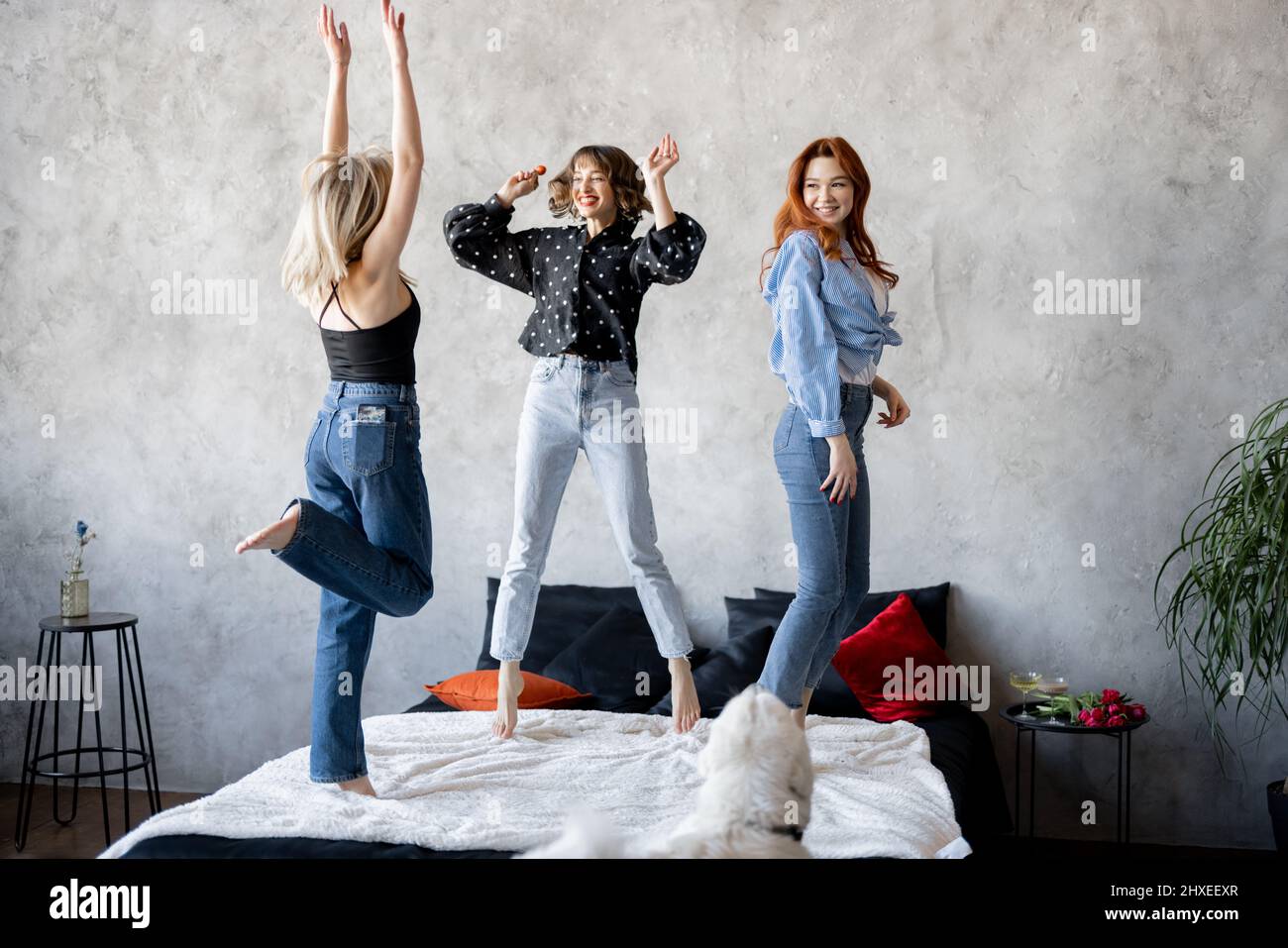 Three young adult girlfriends dressed casual having fun, jumping together on bed. Caucasian women celebrating and having a party at home. Concept of female friendship, beauty and happiness Stock Photo