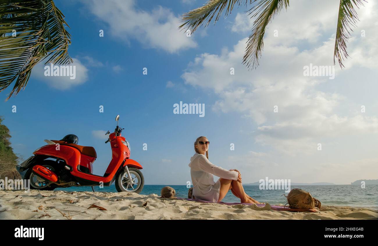 Scooter road trip. Woman alone on red motorbike in white clothes on sand beach by ocean. One girl caucasian tourist walk near tropical palm tree, sea. Stock Photo