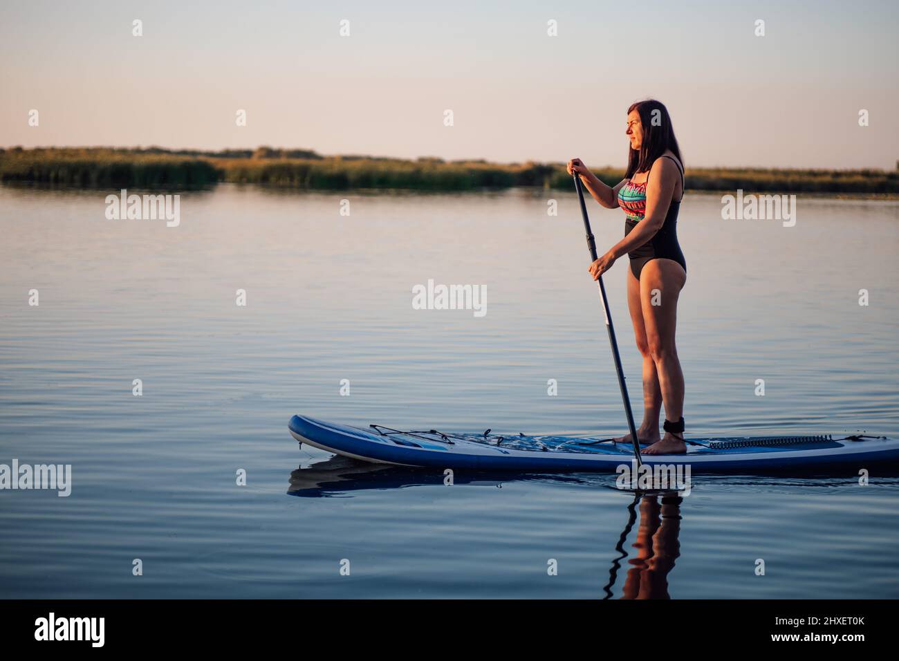 Lady of middle-age balancing on paddle board holding one oar in hands to steer board looking ahead on blue lake wearing swimming dress. Active Stock Photo