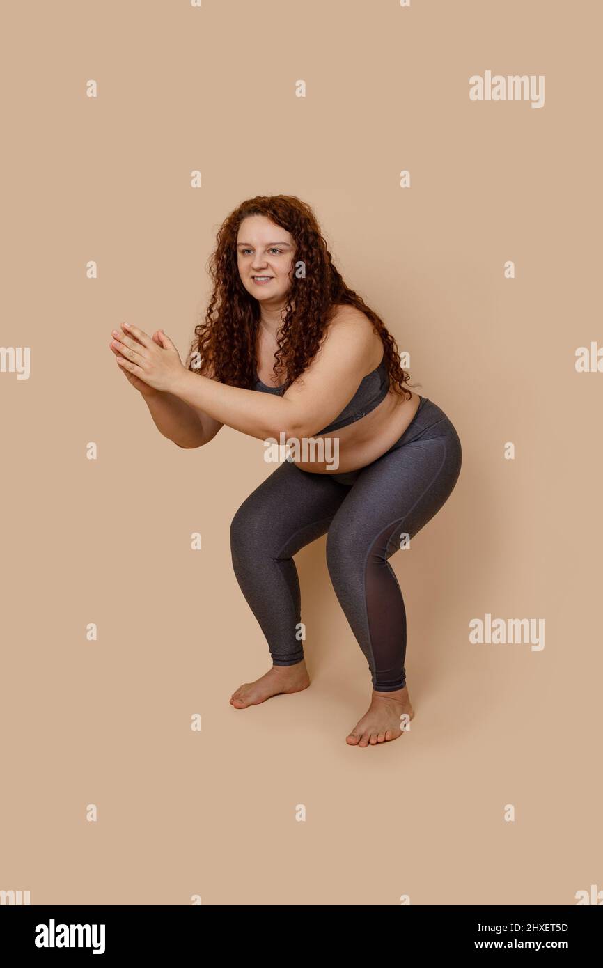 Vertical image of glad ginger massive big obesity fat overweight woman, in gray leggings and top. Knee bending, squat Stock Photo