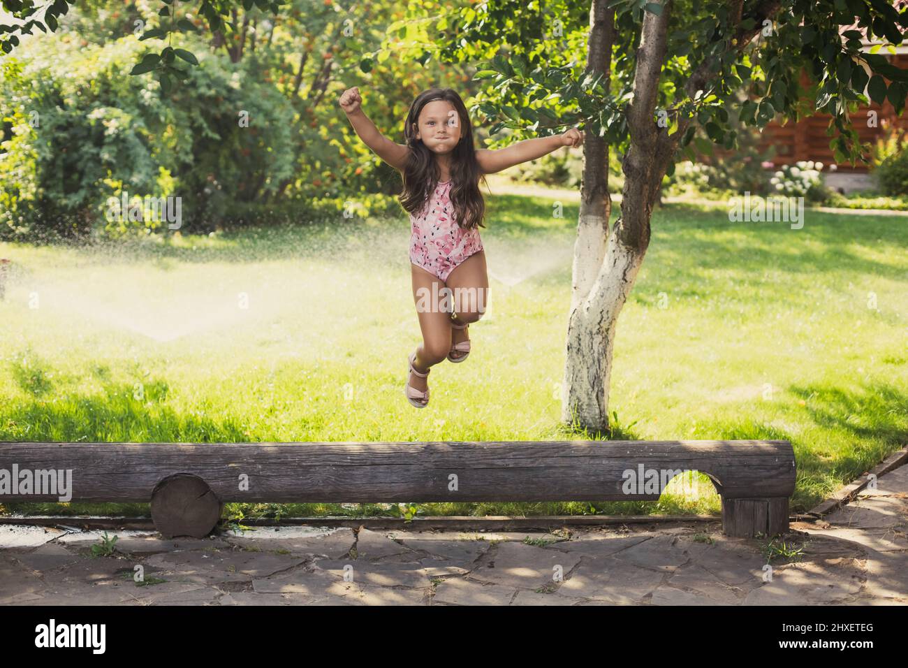 Shining small girl in jump off log smiling with puffy cheeks looking at camera with water sprinklers and greenary in background in daytime. Dreaming Stock Photo