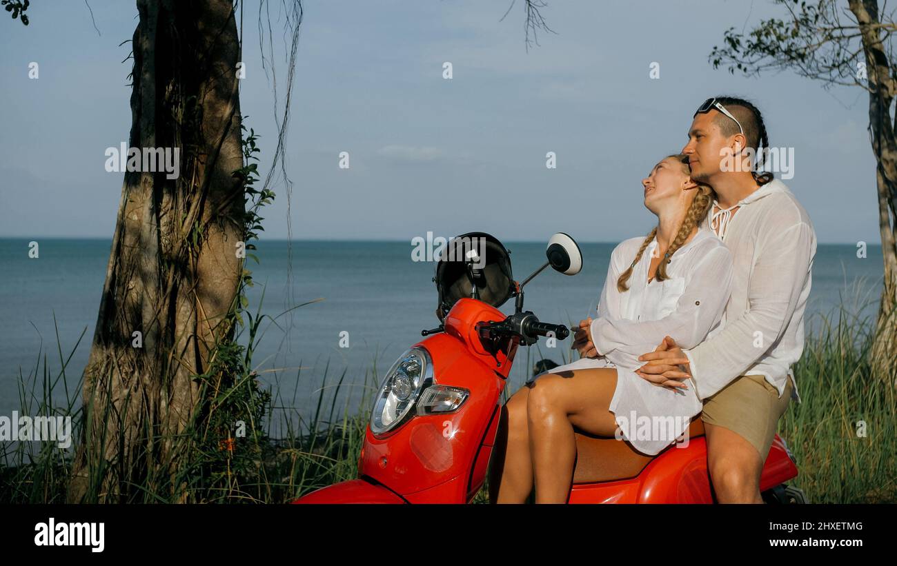 Scooter road trip. Love couple on red motorbike in white clothes on forest road trail. Just married woman and man kiss, hugs, sit on motorbike. Weddin Stock Photo