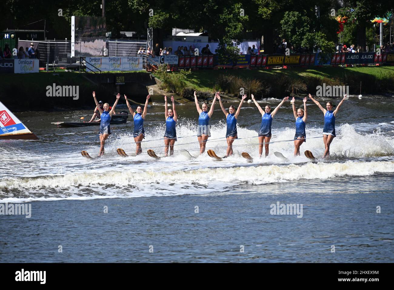 Eight women, in blue costumes, skiing alongside one another with both arms raised at the 2022 Moomba Masters Ski Show Stock Photo