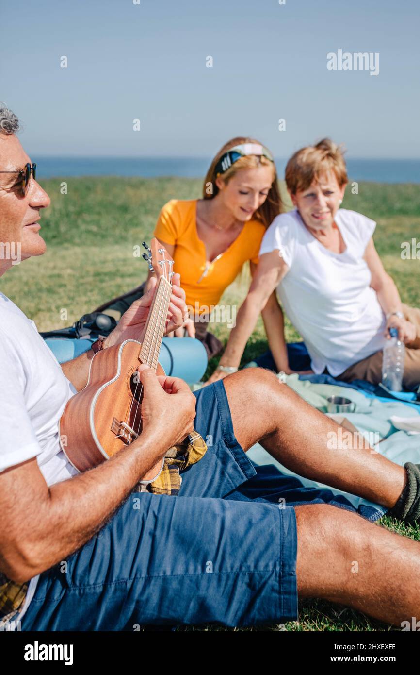 Senior man playing ukulele for his family sitting on a blanket during an excursion Stock Photo