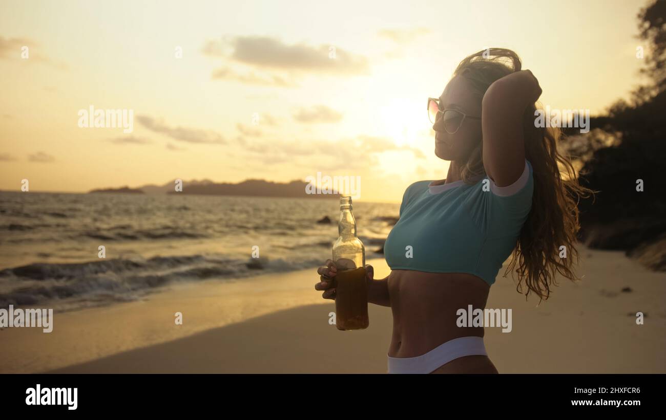 Sexy hot silhouette woman drinking whiskey with orange juice on beach. Rays gold sunset flare through glass. Woman in blue swimsuit, sunglasses. Conce Stock Photo