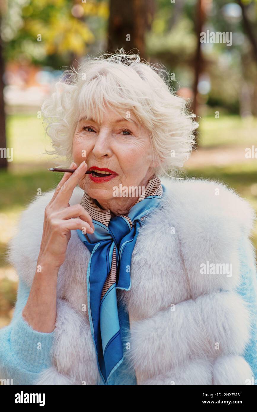 smiling senior elegant stylish fashionable woman with grey hair in fur coat outdoor smoking cigarette. Unhealthy lifestyle, age, positive vibes Stock Photo