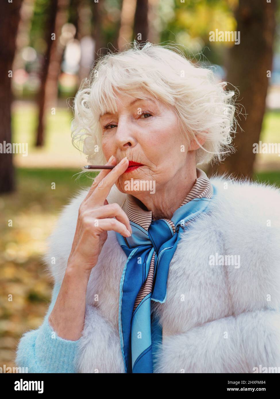 smiling senior elegant stylish fashionable woman with grey hair in fur coat outdoor smoking cigarette. Unhealthy lifestyle, age, positive vibes Stock Photo