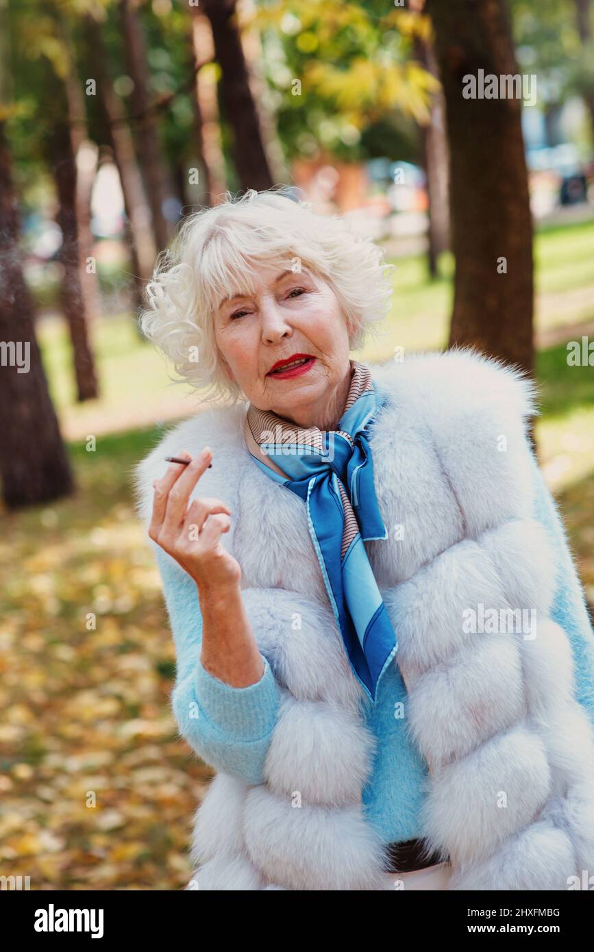 smiling senior elegant stylish fashionable woman with grey hair in fur coat outdoor smoking cigarette. Unhealthy lifestyle, age, positive vibes Stock Photo
