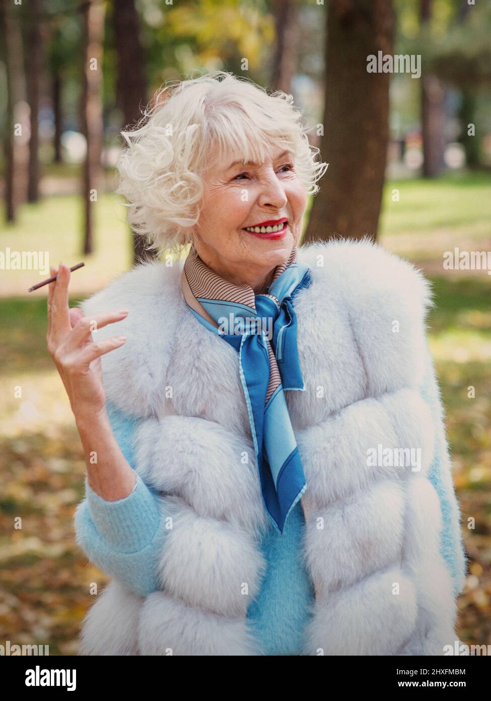 smiling senior elegant stylish fashionable woman with grey hair in fur coat outdoor smoking cigarette. Unhealthy lifestyle, age, positive vibes Stock Photo