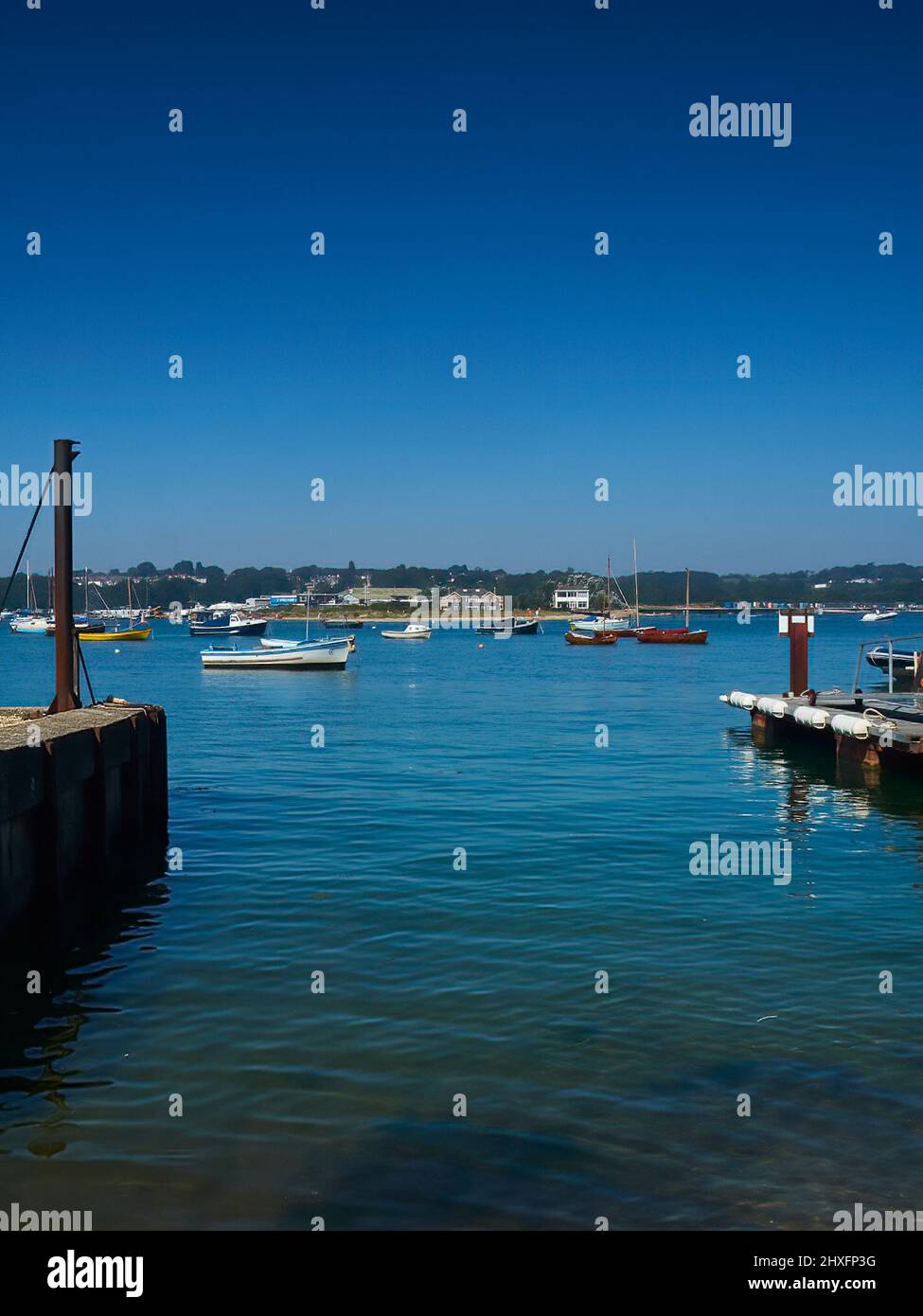 A view from a slipway across Bembridge Harbour, with yachts and boats floating on the mirror-like waters ahead of wooded hills and under blue skies. Stock Photo