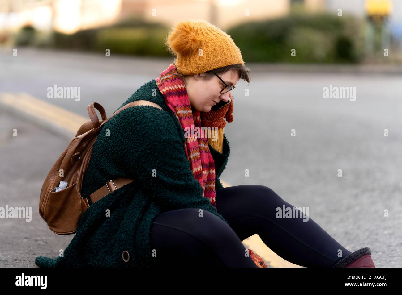 young women 20-25 sat on roadside talks on mobile phone women wears woolly hat and scarf with rucksack on her back Stock Photo