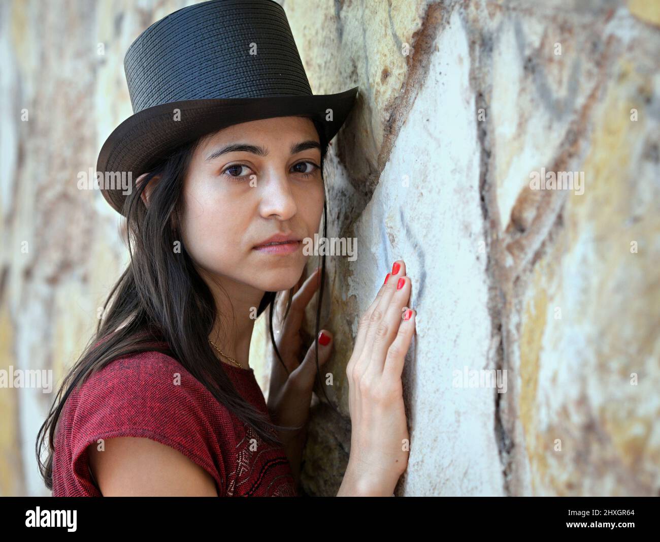 Young beautiful brunette woman with long hair and red fingernails wears a black top hat and looks at the viewer in front of a stone wall background. Stock Photo