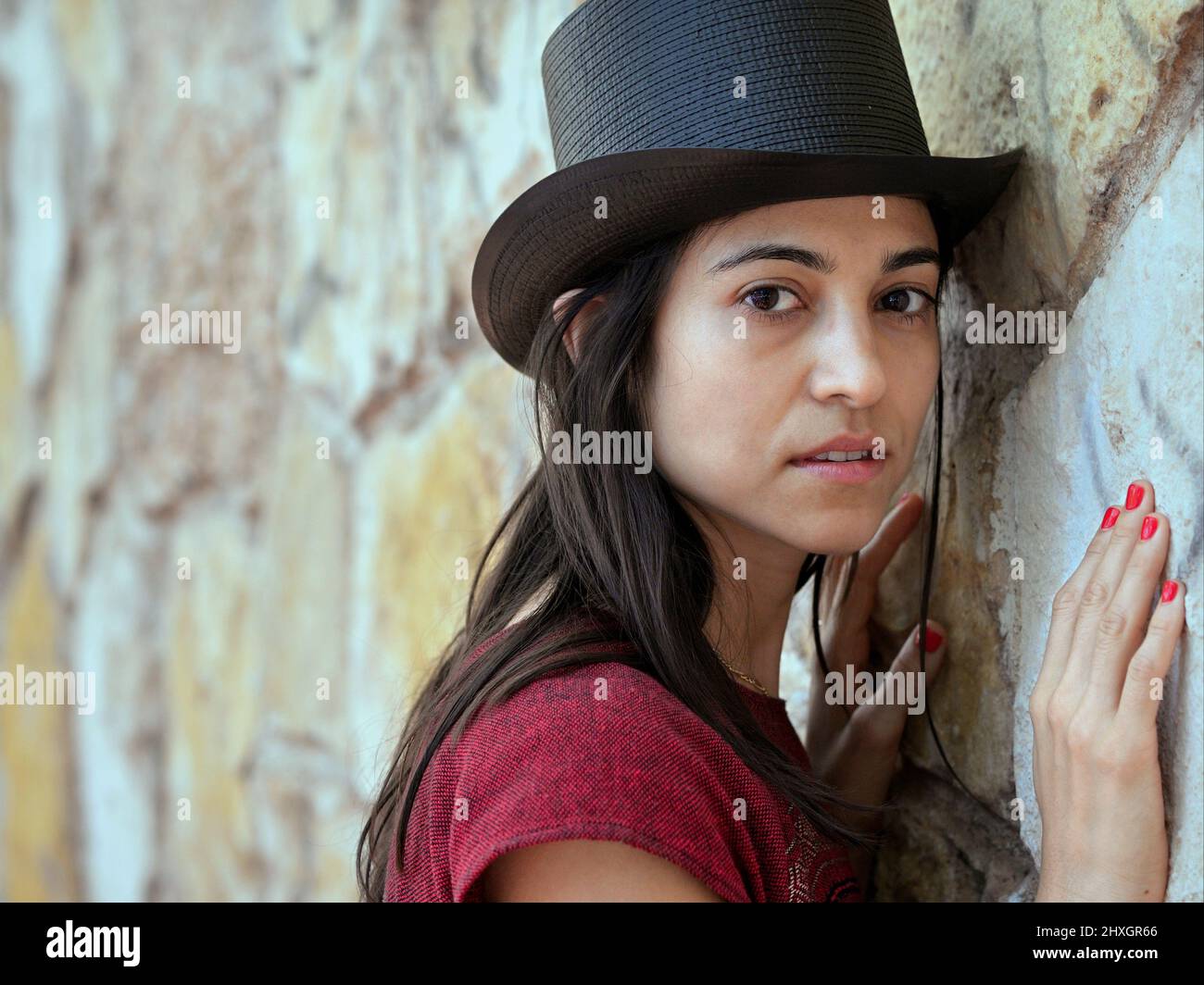 Young beautiful brunette woman with long hair and red fingernails wears a black top hat and looks at the viewer in front of a stone wall background. Stock Photo