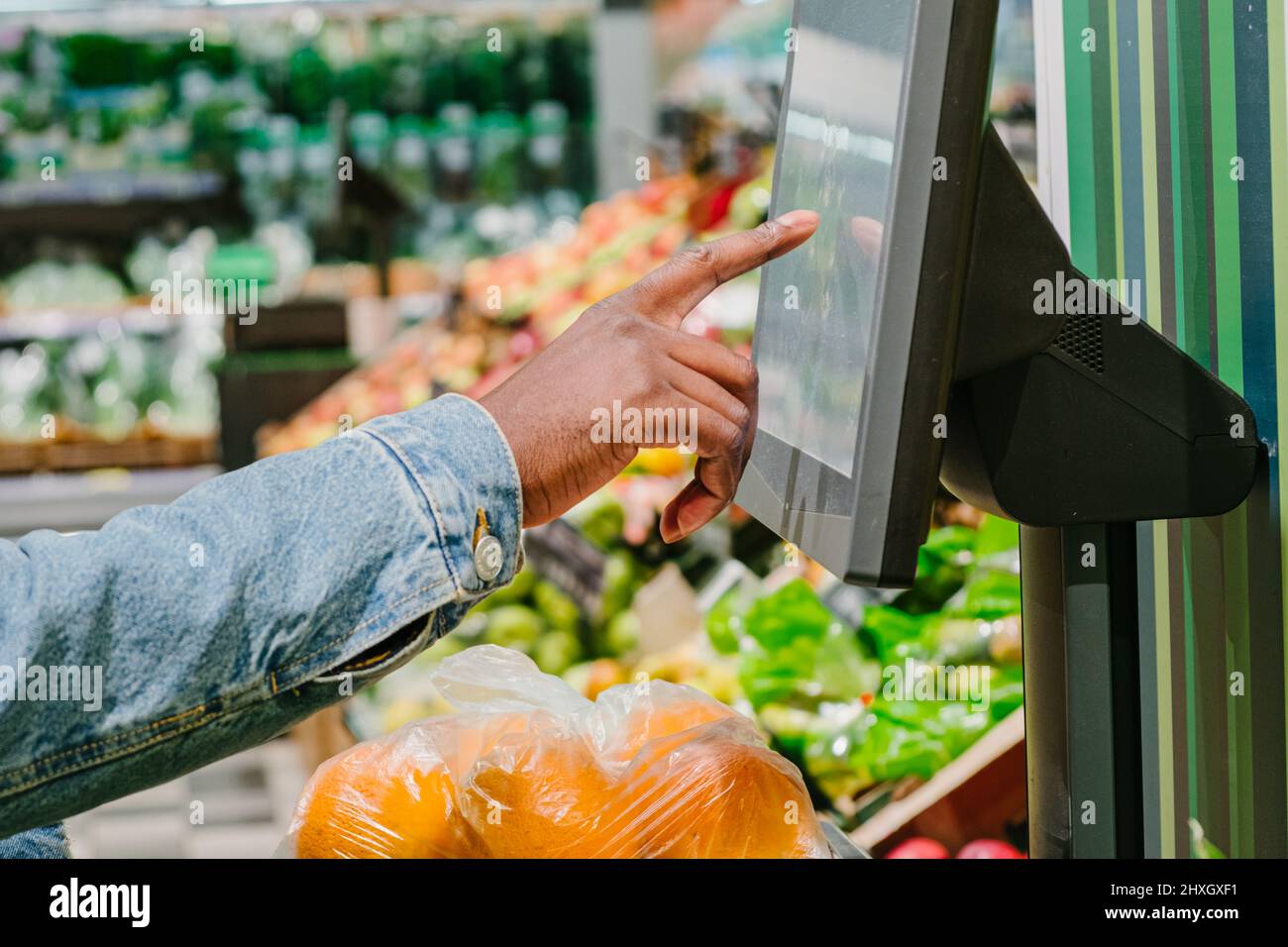 Stylish African-American guy in warm jacket with disposable mask weighs fresh oranges with digital scale in light supermarket Stock Photo