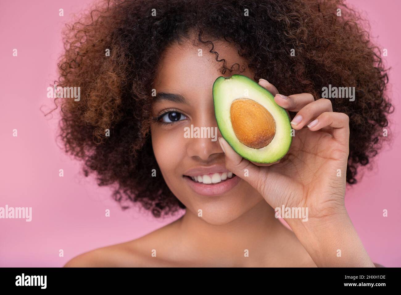 Portrait of a beautiful young dark skinned woman with curly hair covers one eye with half an avocado looks at the camera and smiles on a pink background. Stock Photo