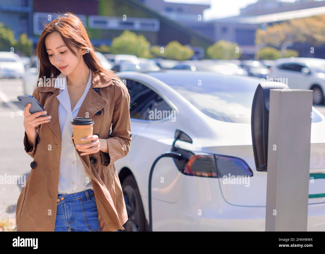 Smiling young woman standing on city parking near electric car, charging automobile battery from small city station, drinking coffee and using smartph Stock Photo
