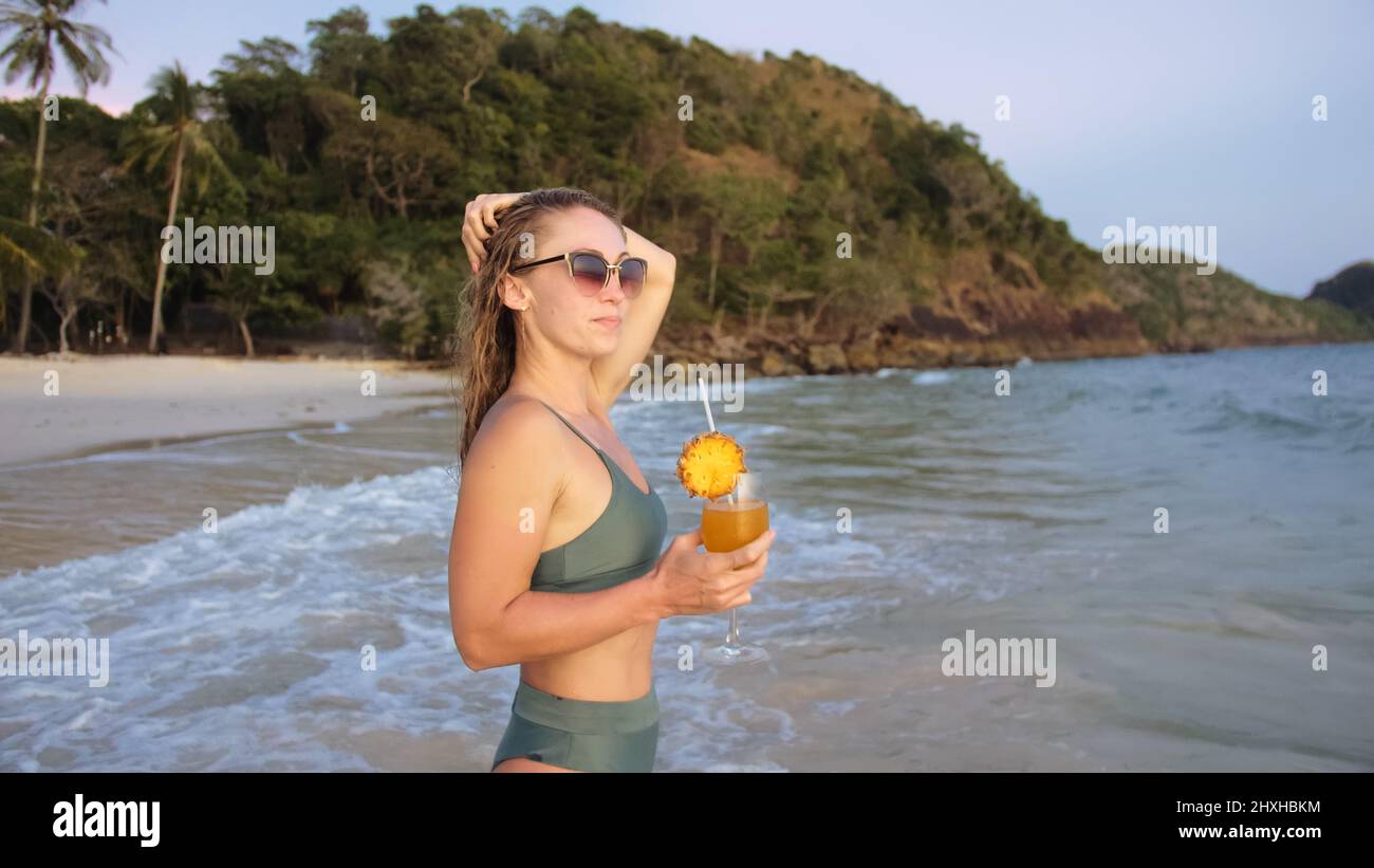 Beautiful woman stand knee-deep in sea on golden sunset. Girl on tropical beach in green swimsuit having fun and waving his hands, drinks her orange c Stock Photo
