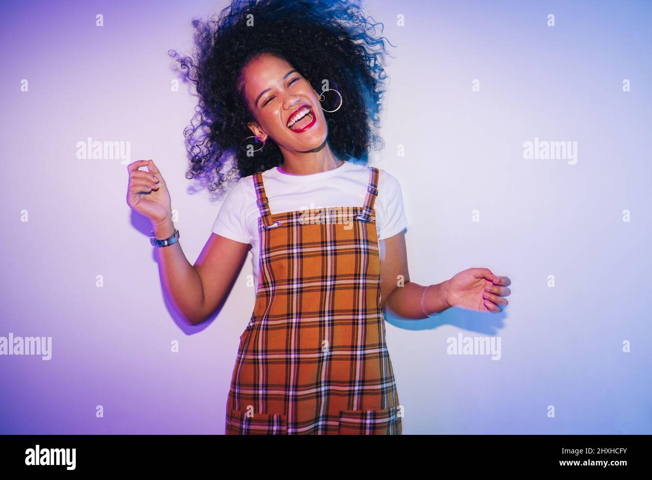 Vibrant young woman dancing and whipping her hair happily. Cheerful young woman laughing joyfully while standing alone in bright neon light. Carefree Stock Photo