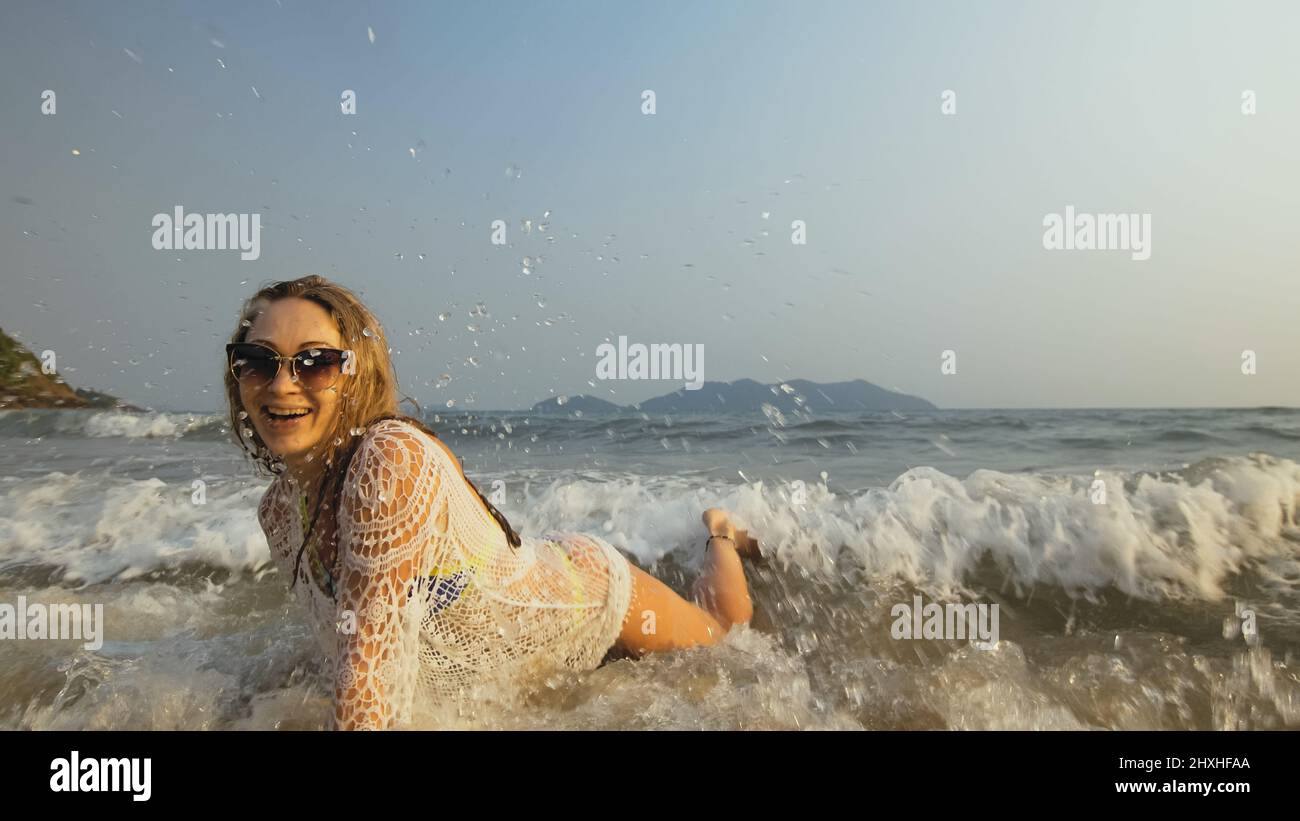 Sexy fit woman in a white tunic on the beach, near the stormy sea. Happy playful blonde with wet curly hair, in sunglasses. Lady tourist enjoying vaca Stock Photo