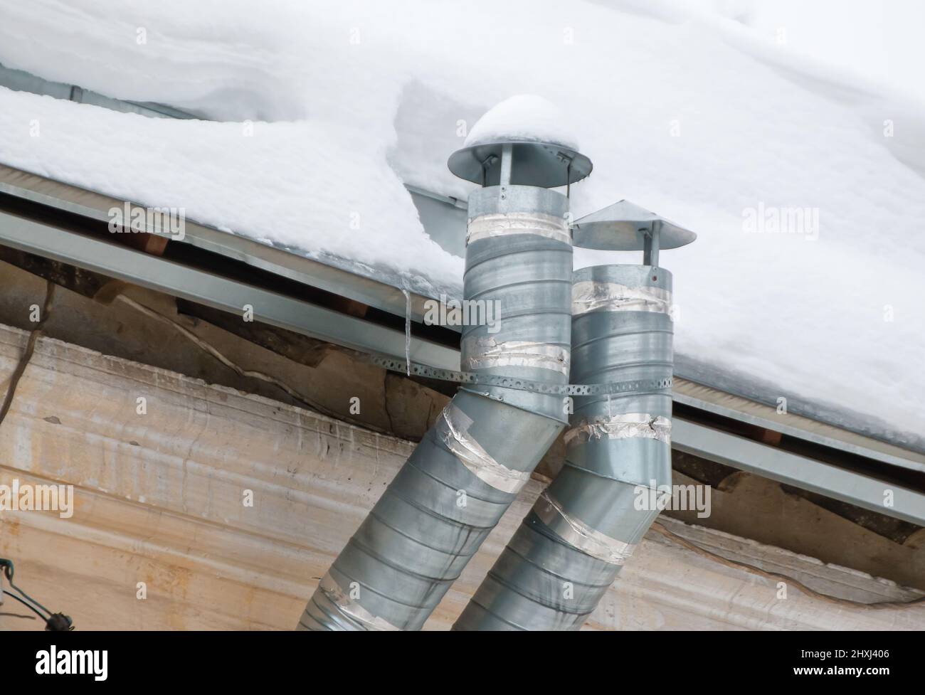 Two metal exhaust pipes on the roof of the house. There is white snow on the surface. Against the background of a gray sky. Cloudy winter day, soft light. Stock Photo