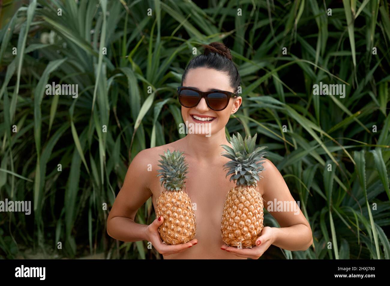 Beautiful cheerful nude girl holding a pineapple at the breast on the tropical background . Woman with bronze tan in bikini. Covering her breast with Stock Photo