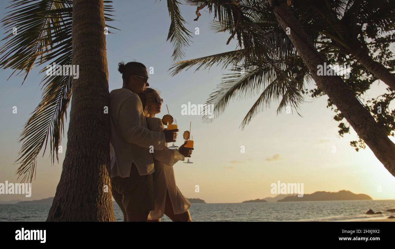 The man hugs a woman. Cute loving couple in white shirt and sunglasses, near palm tree, kissing, hugging, relax and drinking cocktail. Concept tropica Stock Photo