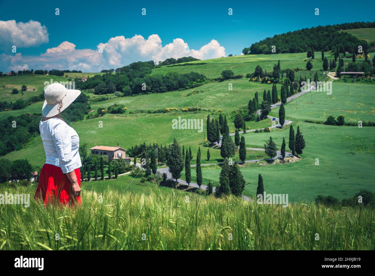 Pretty cheerful woman in a red skirt white shirt and a straw hat enjoying the view in Tuscany. Famous rural touristic place with grain fields and curv Stock Photo