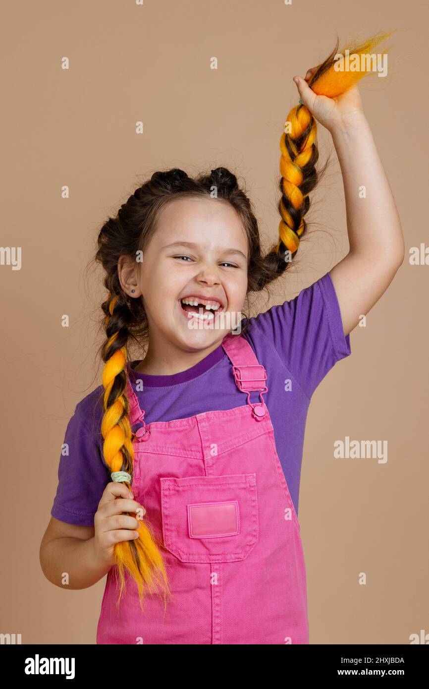 Laughing happy, young girl with yellow kanekalon braids with opened mouth with missing tooth holding hair looking at camera wearing pink jumpsuit and Stock Photo