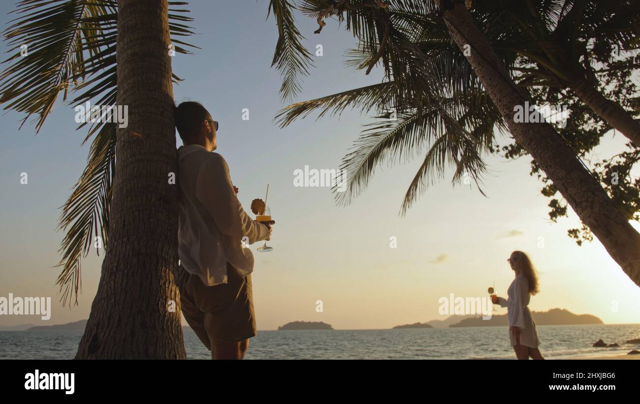 The woman goes to the man. Loving couple in white shirt and sunglasses, near palm tree, kissing, hugging, relax and drinking cocktail. Concept tropica Stock Photo