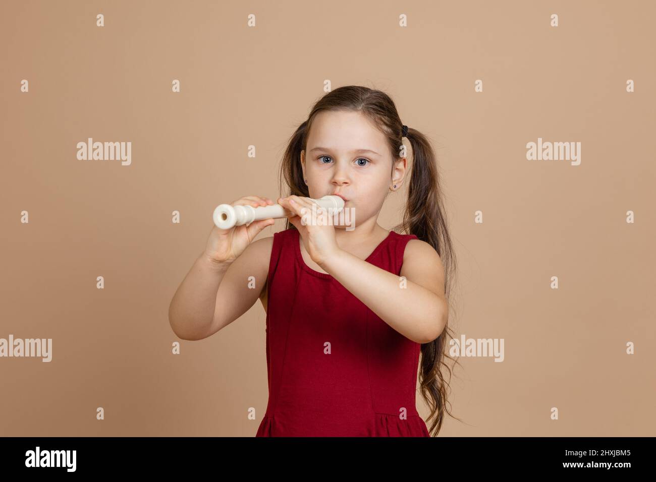 Girl in red dress thoughtfully play melody on flute, blowing air into duct, beige background. Learn to play woodwind musical instrument. Stock Photo