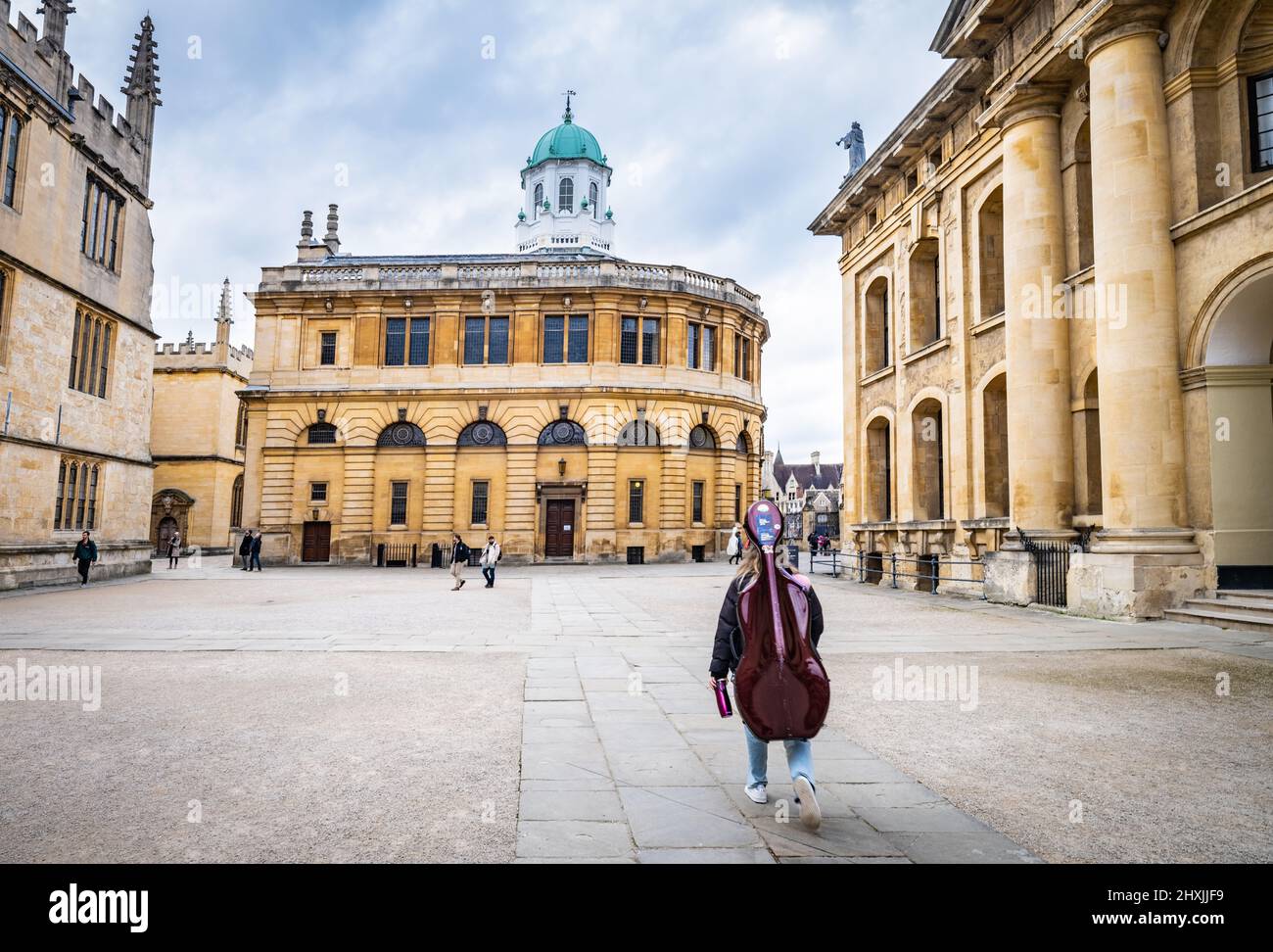 A  young musician  carrying her cello through Bodleian Library courtyard in Oxford, UK.. The Old Bodleian Library (left), Sheldonian Theatre (centre) Stock Photo
