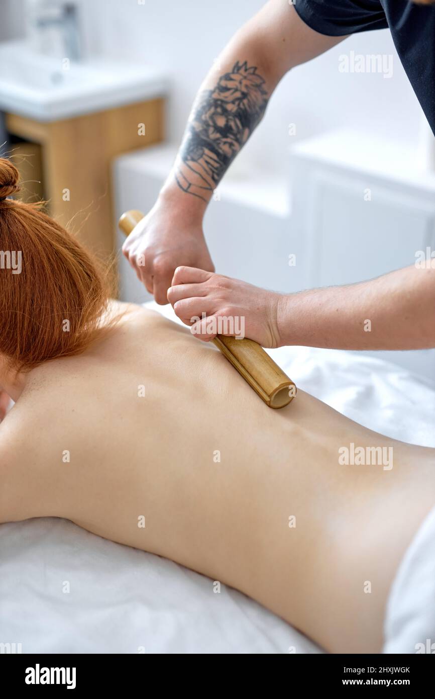Bamboo massage. unrecognizable male masseur massages the body using bamboo sticks in wellness center. cropped caucasian european lady client lying nak Stock Photo