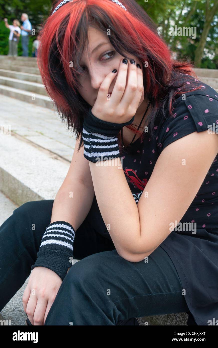 Punk emo girl, young adult with black red hair, sitting on a staircase outdoors, holding her head, looking away, horizontal Stock Photo