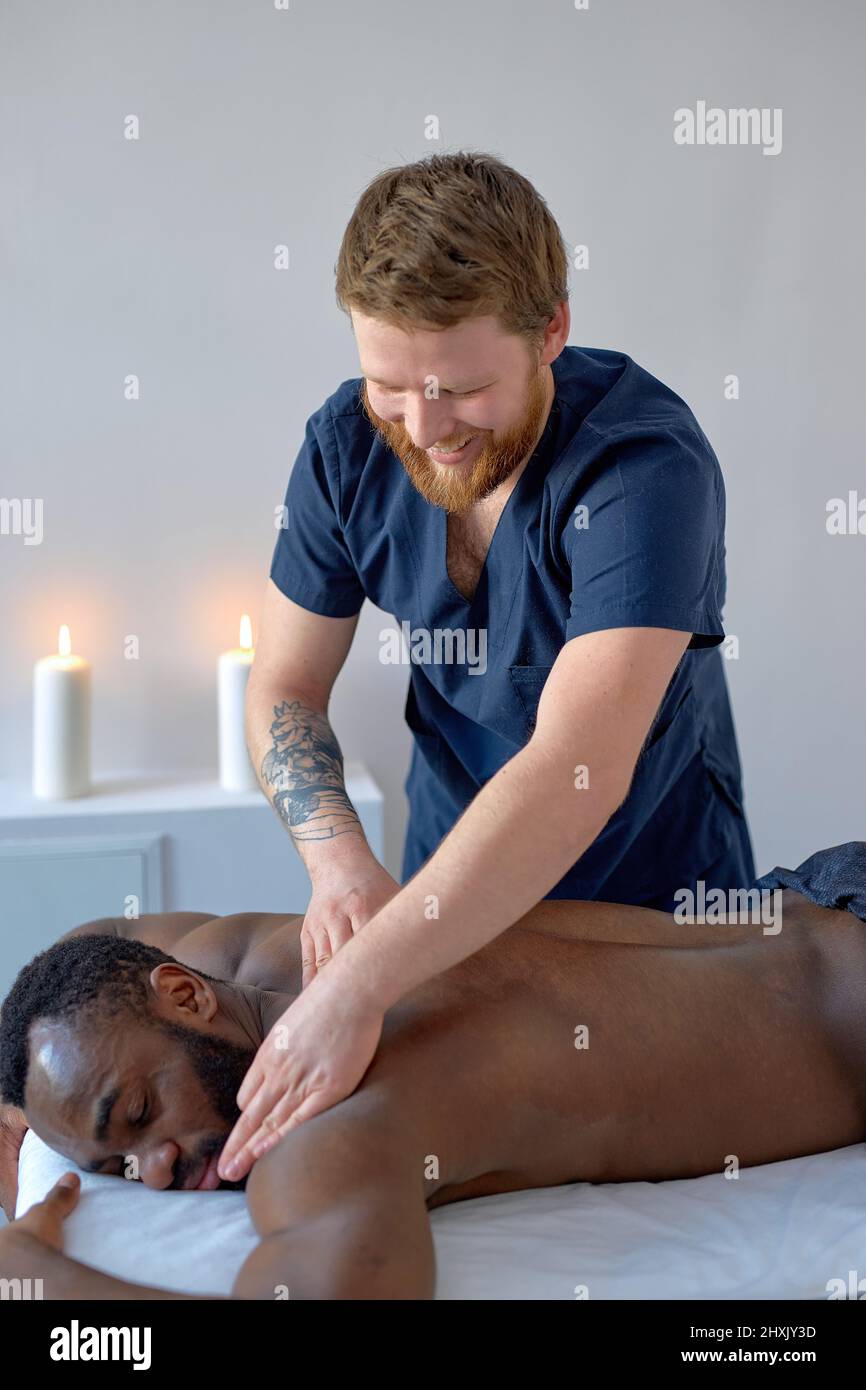 confident caucasian male masseur with strong hands professionally massaging scapulas and shoulders of male. handsome naked young black man with perfec Stock Photo