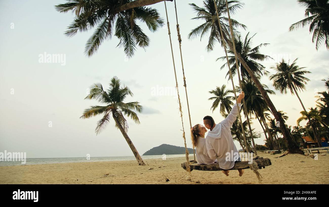 Cute loving couple on the swing on tropical beach. Woman and man in white shirt enjoy gold sunset and life in beaches, palm tree. Evening warm sunset. Stock Photo