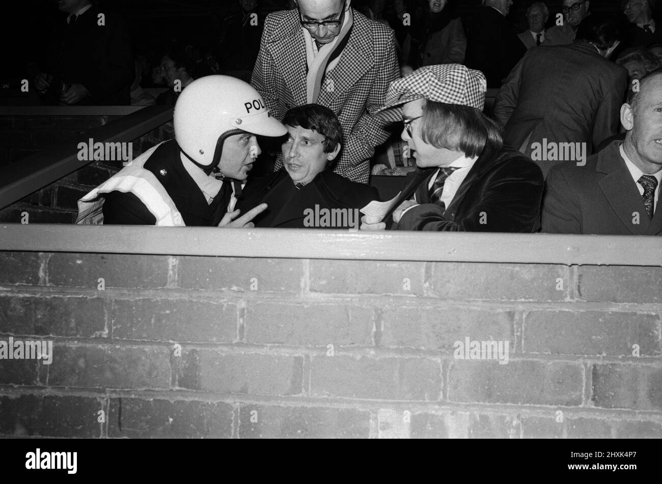 A policeman talking to Graham Taylor and Elton John, who are watching the football match, West Bromwich Albion v Watford.  25 October 1977. Stock Photo