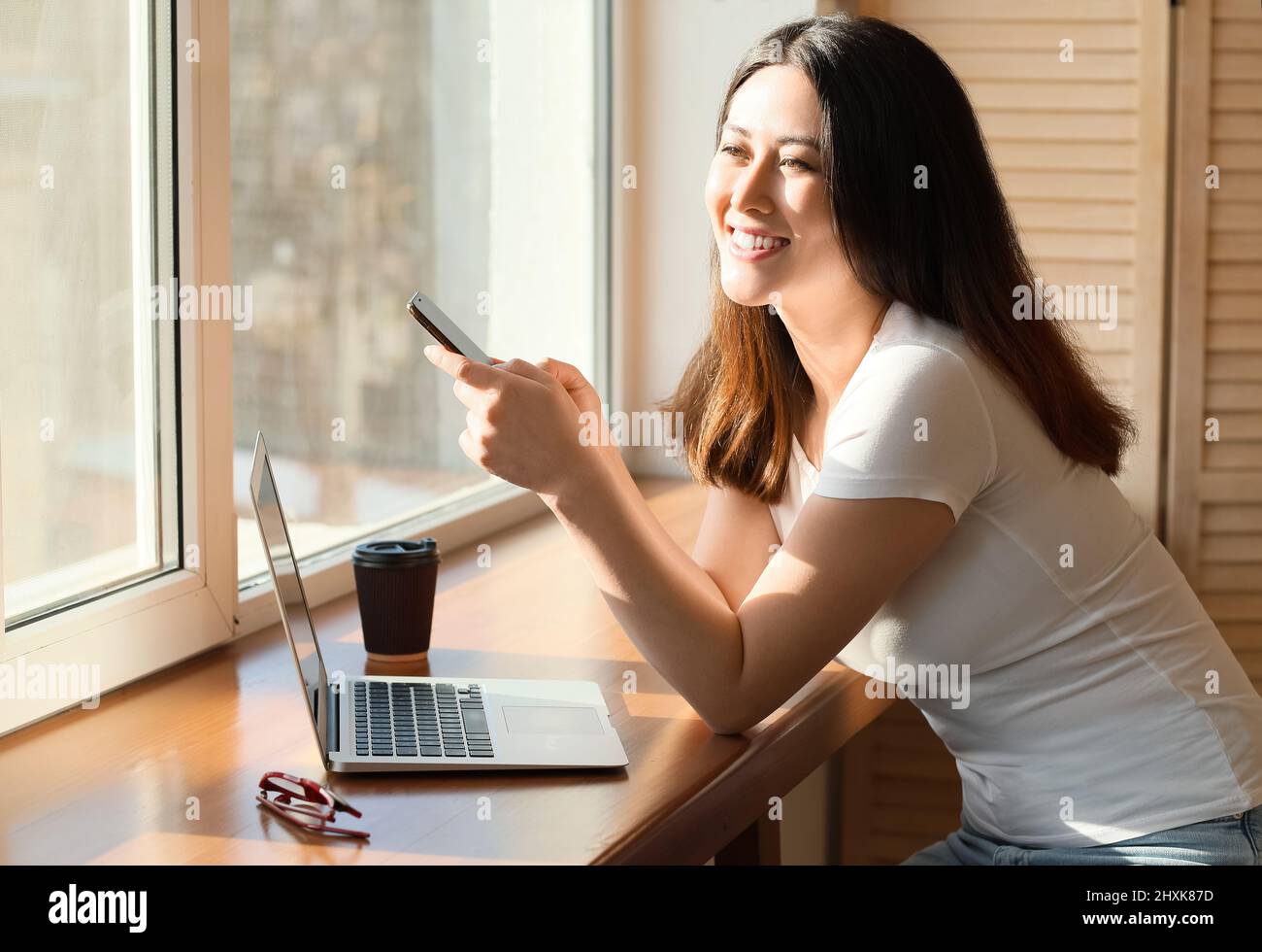 Beautiful young Asian woman with mobile phone and laptop near window ...