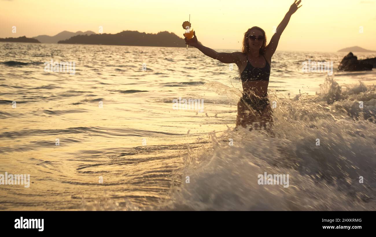 Silhouette glow cinematic view. Sexy woman stand knee-deep in sea on golden sunset. Girl on tropical beach in green swimsuit having fun and waving his Stock Photo