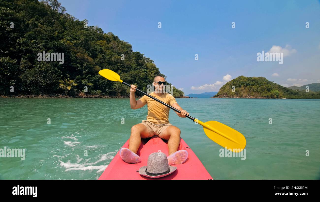Young man with sunglasses and hat rows pink plastic canoe along sea against green hilly islands with wild jungles. Traveling to tropical countries. Strong guy is sailing on kayak in ocean, front view. Stock Photo