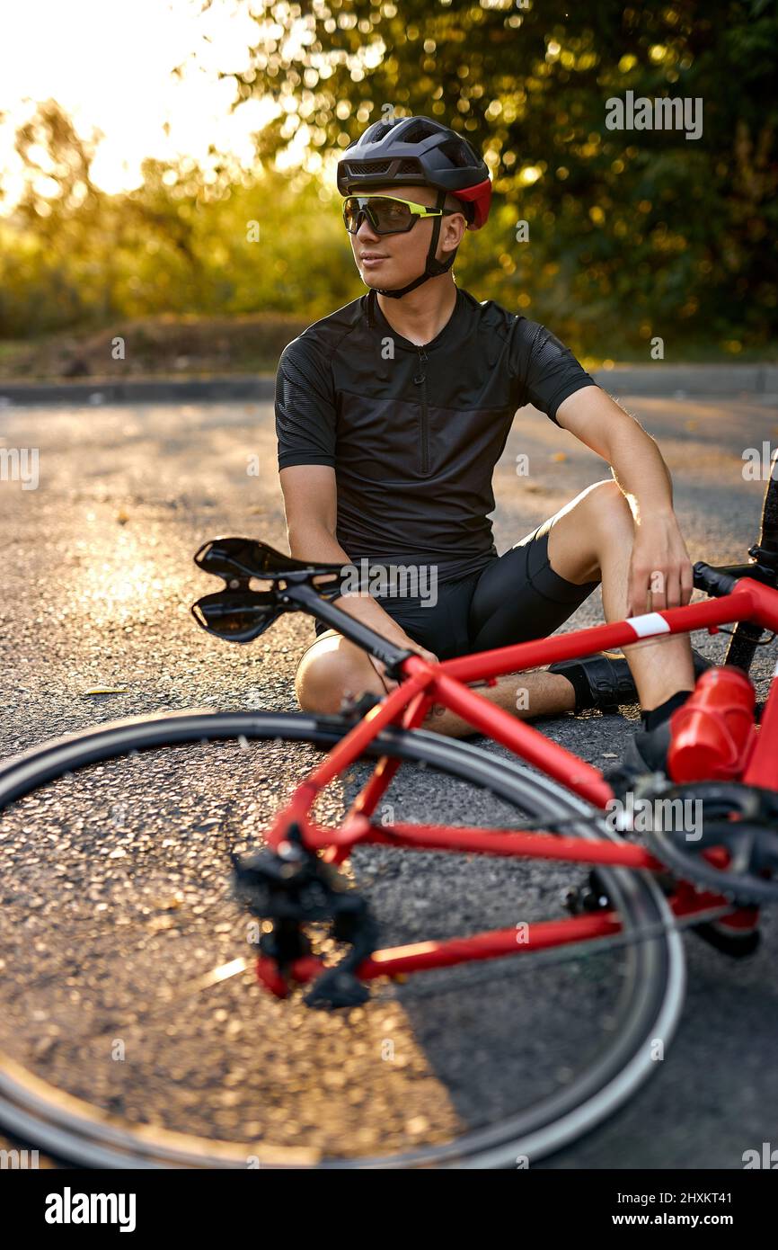Sporty guy biker in cycling clothes and helmet sit on road near bicycle, having rest. Man resting and relaxing after training on bicycle in nature. He Stock Photo