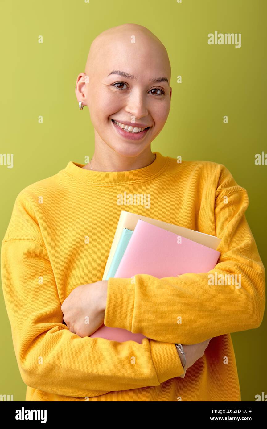 Excited female with bald head holding books in hands and smiling, enjoying education, studying. Hairless lady in casual yellow shirt posing at camera, Stock Photo