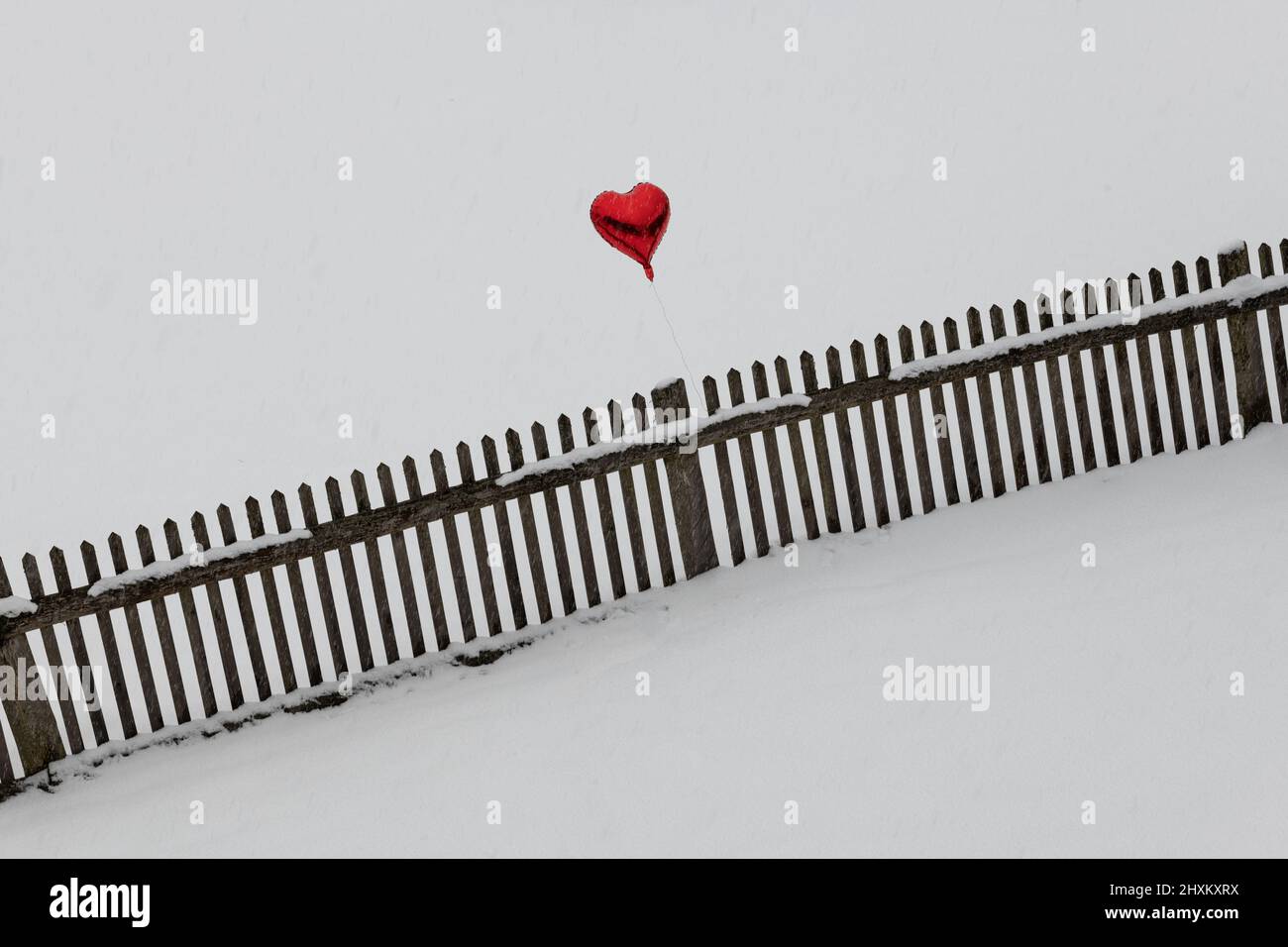 heart shaped red balloon hovering over wooden fence in snow covered llandscape Stock Photo
