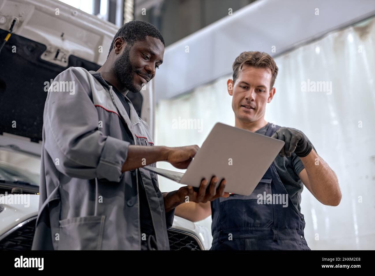 team of diverse mechanics in uniform examining car, using laptop while working in auto service. black and caucasian Auto mechanics checking car,servic Stock Photo
