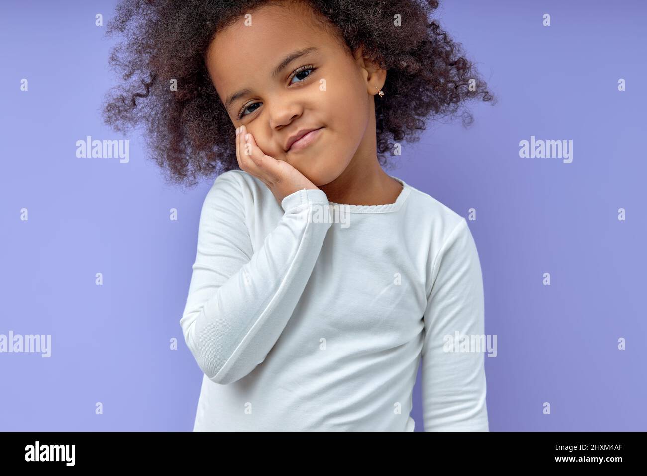adorable young girl kid keeps hands on cheeks,smiles satisfied,amiably, has fun chatting with friends, wears casual clothes, isolated over purple back Stock Photo