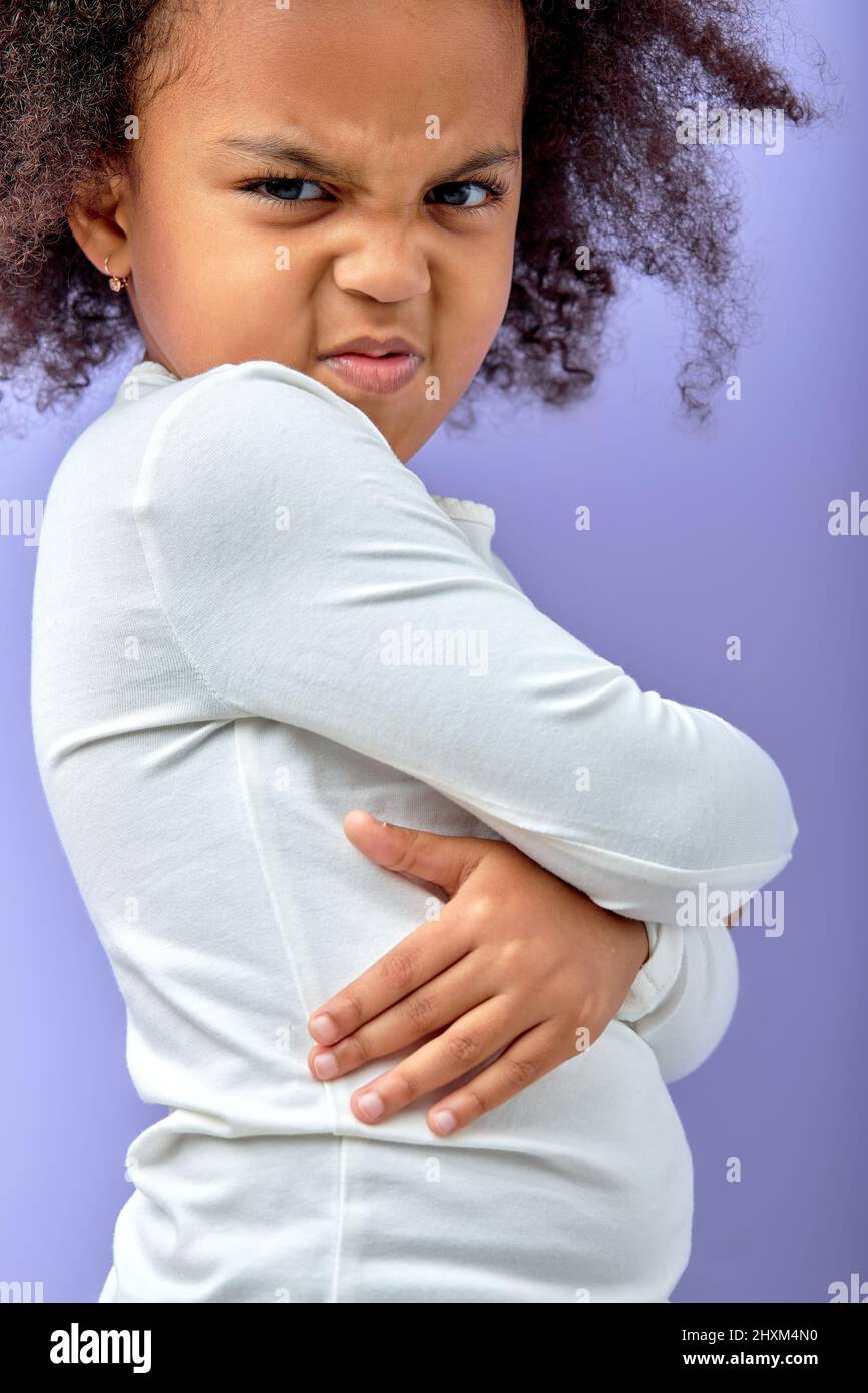 Naughty child. Disobedience problem. Discipline punishment. Portrait of angry black offended little girl in white with crossed arms isolated on purple Stock Photo